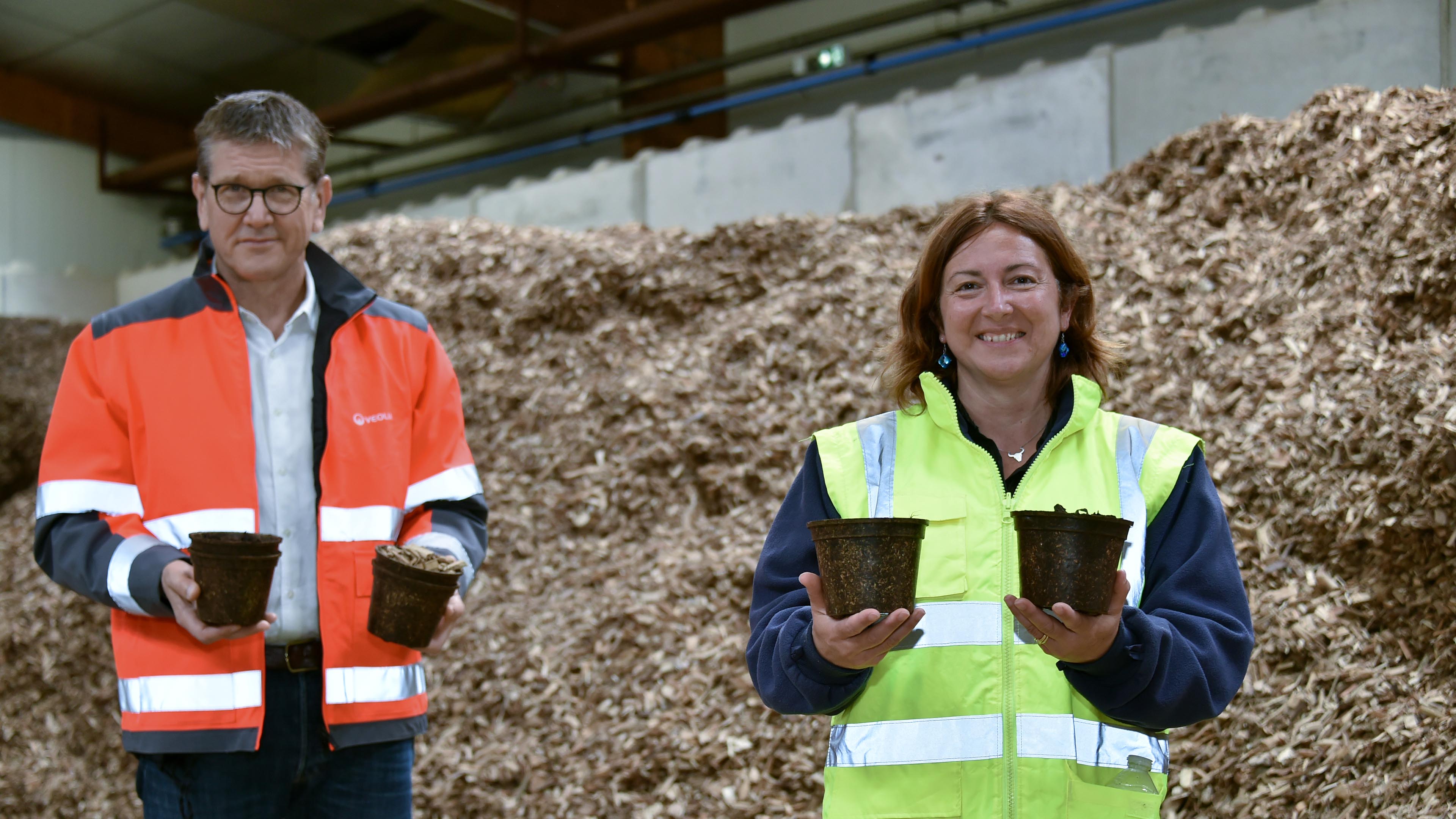 Deux personnes posent avec des pots de l'entreprise, devant un tas de déchets de bois